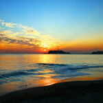 夕暮れの海に静かな波が寄せる中、遠くの小さな島と塔が淡く浮かぶ情景｜懐かしい情景が静かによみがえる、やさしい記憶のイメージ写真