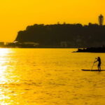 夕暮れの海辺で静かにパドルボードを漕ぐ人のシルエット｜懐かしい情景が静かによみがえる、やさしい記憶のイメージ写真