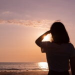 夕暮れの海辺で女性が片手をかざして夕日を見つめるシルエットの情景｜懐かしい情景が静かによみがえる、やさしい記憶のイメージ写真