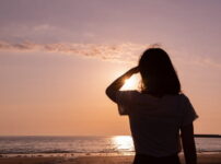 夕暮れの海辺で女性が片手をかざして夕日を見つめるシルエットの情景｜懐かしい情景が静かによみがえる、やさしい記憶のイメージ写真