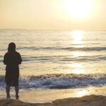 【夕暮れの海辺に立つ人影と、波に反射する柔らかな光の情景】｜懐かしい情景が静かによみがえる、やさしい記憶のイメージ写真