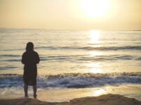【夕暮れの海辺に立つ人影と、波に反射する柔らかな光の情景】｜懐かしい情景が静かによみがえる、やさしい記憶のイメージ写真