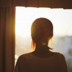 窓辺で柔らかな光に包まれながら外を見つめる女性の後ろ姿｜懐かしい情景が静かによみがえる、やさしい記憶のイメージ写真