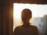 窓辺で柔らかな光に包まれながら外を見つめる女性の後ろ姿｜懐かしい情景が静かによみがえる、やさしい記憶のイメージ写真