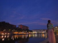 夕暮れの水辺で都会の灯りを静かに見つめる女性の後ろ姿｜懐かしい情景が静かによみがえる、やさしい記憶のイメージ写真