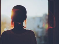 夕暮れの窓辺で夕日を見つめる女性の後ろ姿と柔らかな光｜懐かしい情景が静かによみがえる、やさしい記憶のイメージ写真