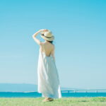 夏の海辺で風に揺れる白いワンピースの女性が、水平線を見つめて立つ後ろ姿の情景｜懐かしい情景が静かによみがえる、やさしい記憶のイメージ写真