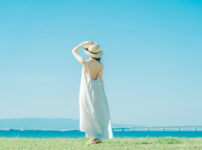 夏の海辺で風に揺れる白いワンピースの女性が、水平線を見つめて立つ後ろ姿の情景｜懐かしい情景が静かによみがえる、やさしい記憶のイメージ写真
