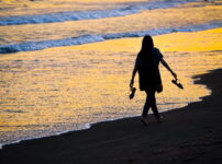 夕暮れの海辺を、サンダルを手にそっと歩く人物のシルエットが柔らかい光に染まる情景｜懐かしい情景が静かによみがえる、やさしい記憶のイメージ写真